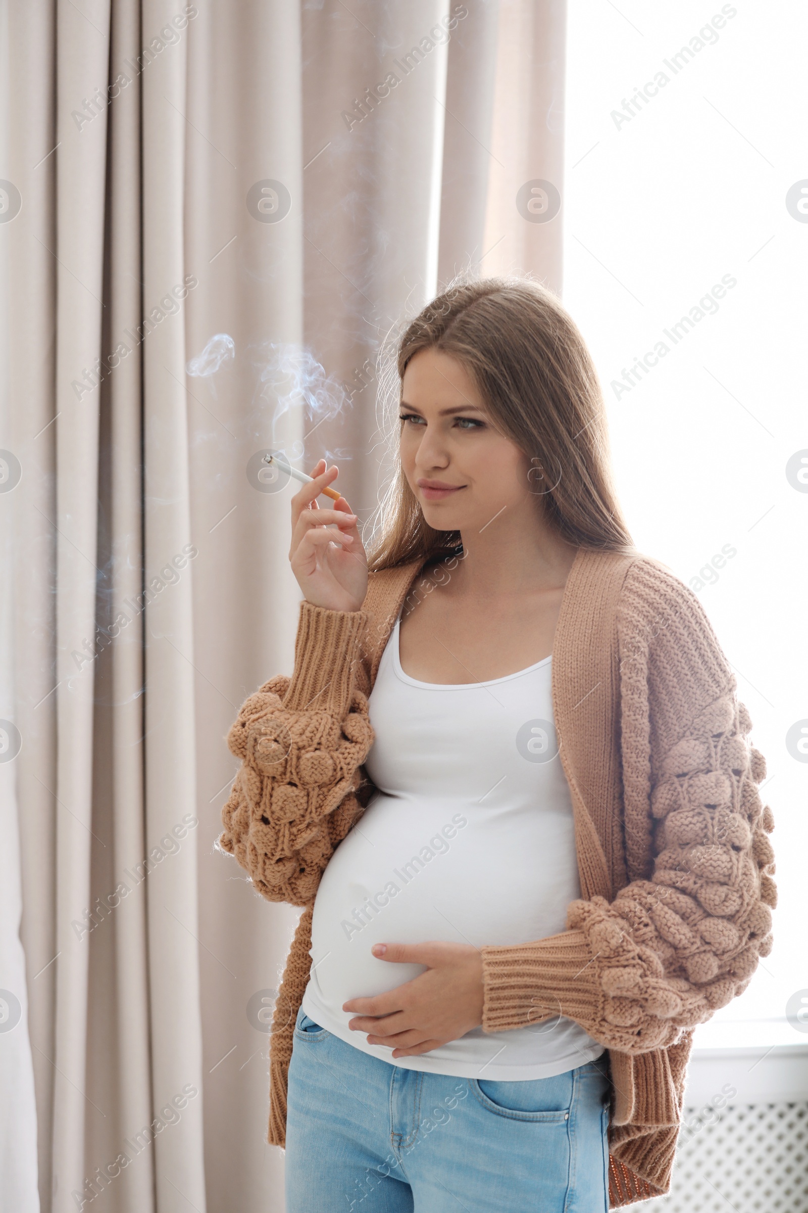 Photo of Young pregnant woman smoking cigarette at home. Harm to unborn baby