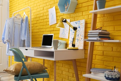 Photo of Fashion designer's workplace with wooden furniture and laptop near yellow brick wall. Stylish interior