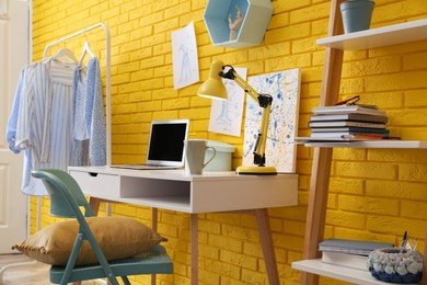 Fashion designer's workplace with wooden furniture and laptop near yellow brick wall. Stylish interior