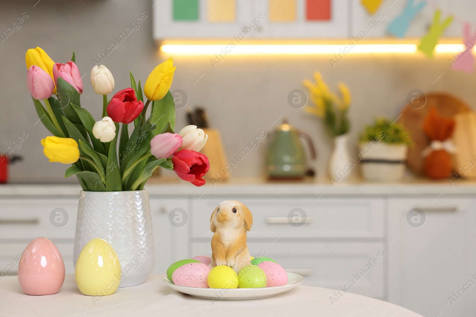 Photo of Bouquet of tulips, painted eggs and Easter decorations on white table in kitchen. Space for text