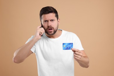 Photo of Emotional man with credit card talking on smartphone against beige background. Be careful - fraud