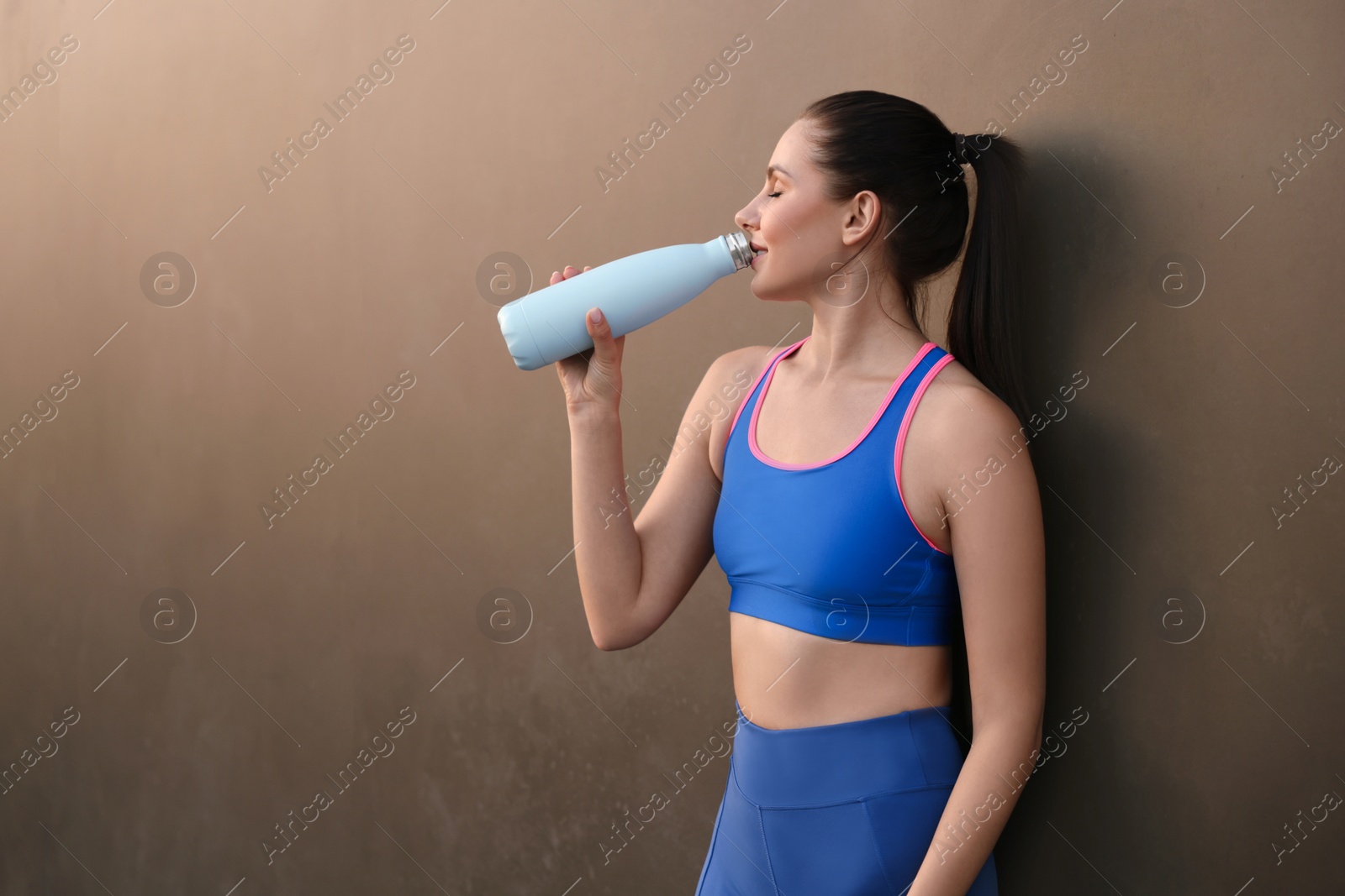 Photo of Beautiful woman in stylish sportswear drinking water near black wall, space for text