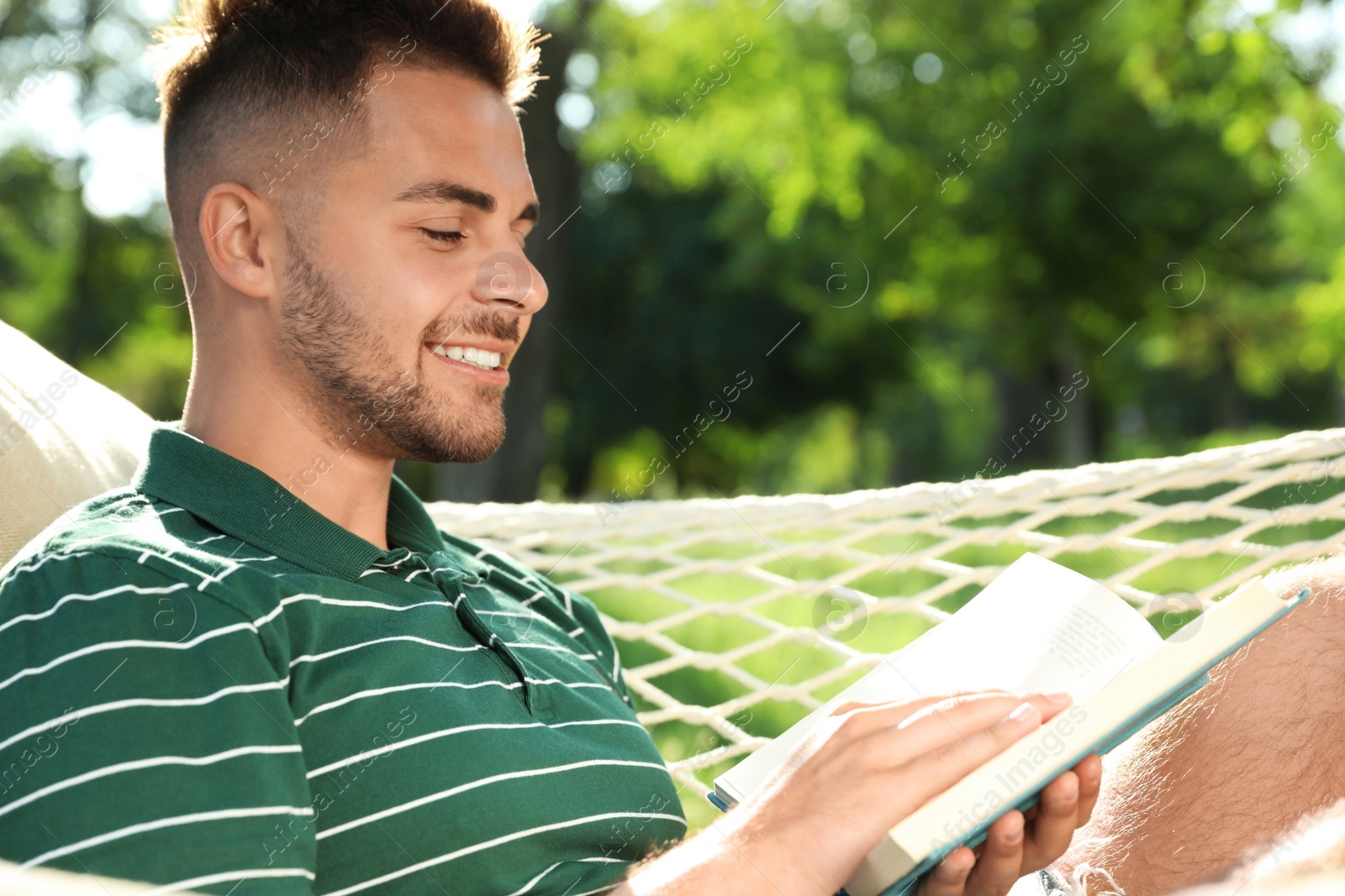 Photo of Young man reading book in comfortable hammock at green garden, closeup