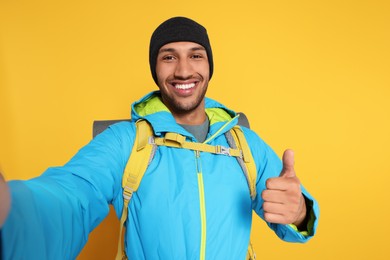 Photo of Happy tourist with backpack taking selfie and showing thumb up on yellow background