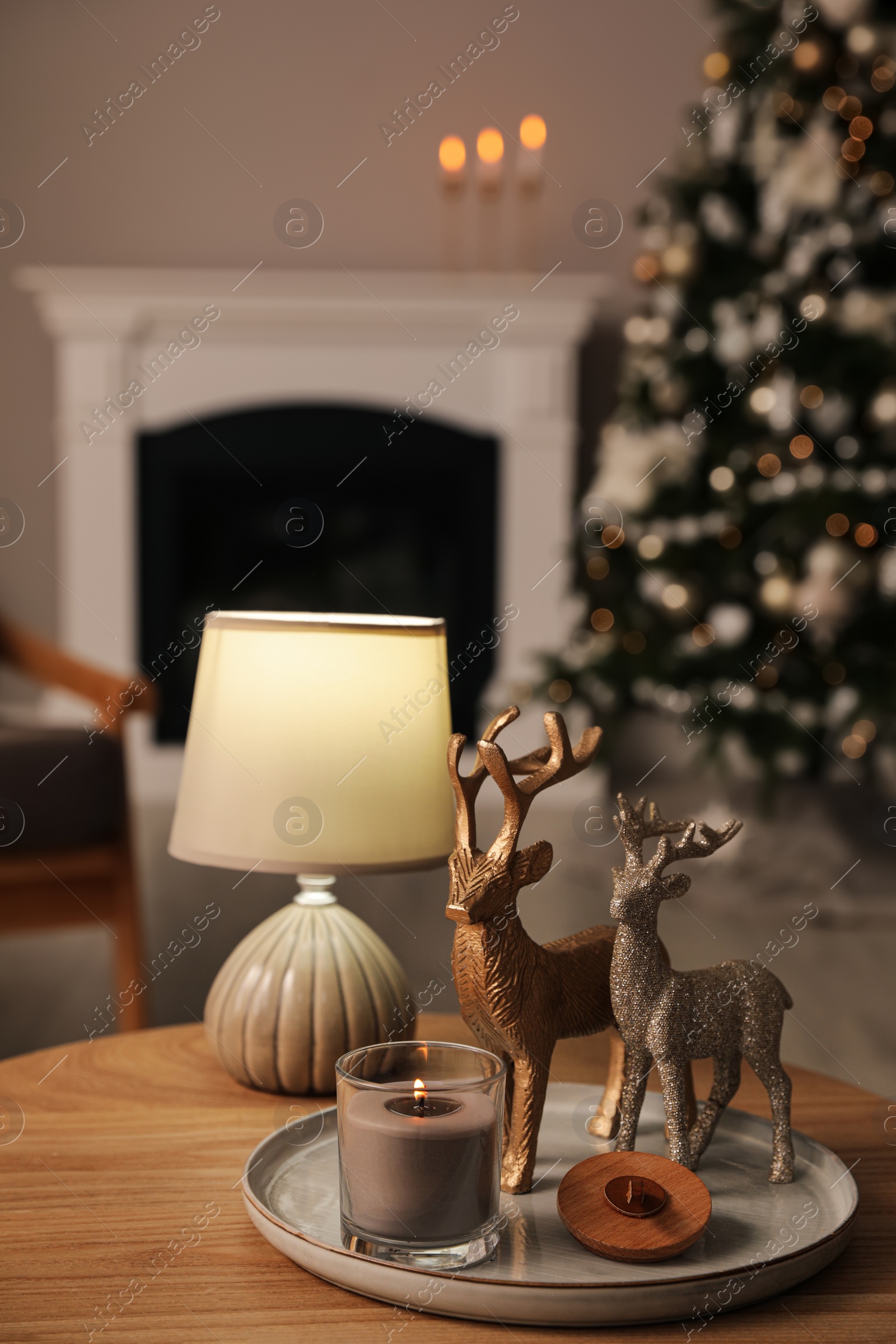 Photo of Deer figures, burning candle with lamp on wooden table and Christmas tree in living room