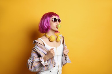 Photo of Fashionable young woman in colorful wig with headphones blowing bubblegum on yellow background, space for text