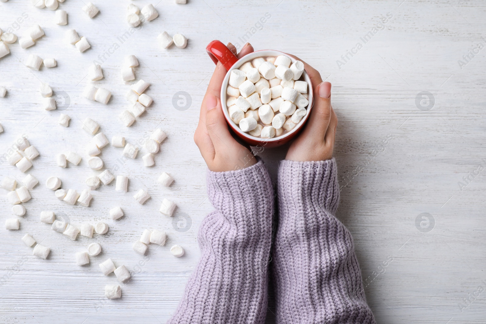 Photo of Woman holding cup of tasty cocoa with marshmallows on white wooden table, top view