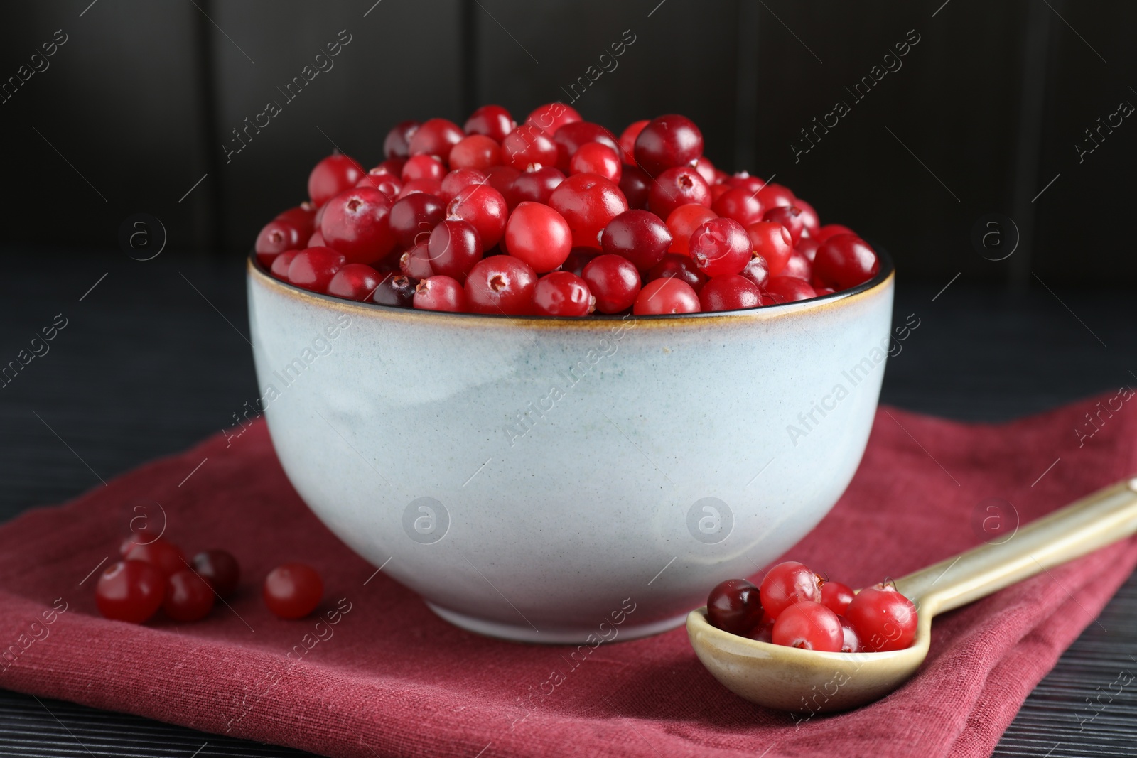 Photo of Cranberries in bowl and spoon on black wooden table, closeup