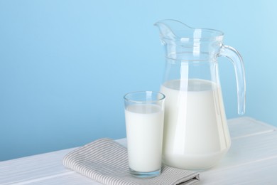 Photo of Jug and glass of fresh milk on white wooden table against light blue background, space for text