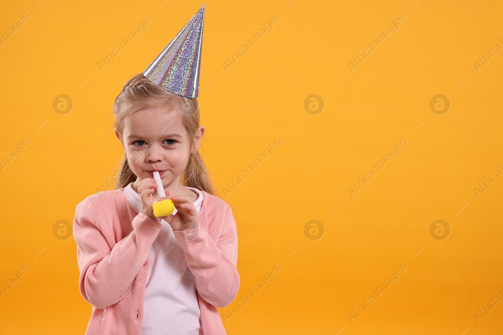 Photo of Birthday celebration. Cute little girl in party hat with blower on orange background, space for text