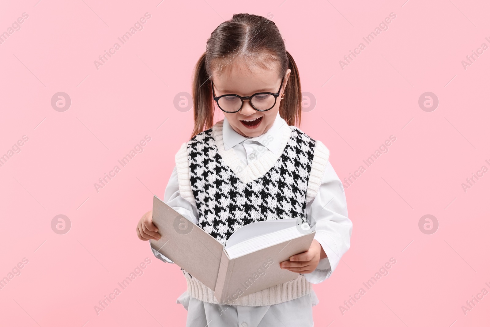 Photo of Cute little girl in glasses reading book on pink background