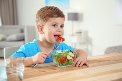 Photo of Adorable little boy eating vegetable salad at table in room