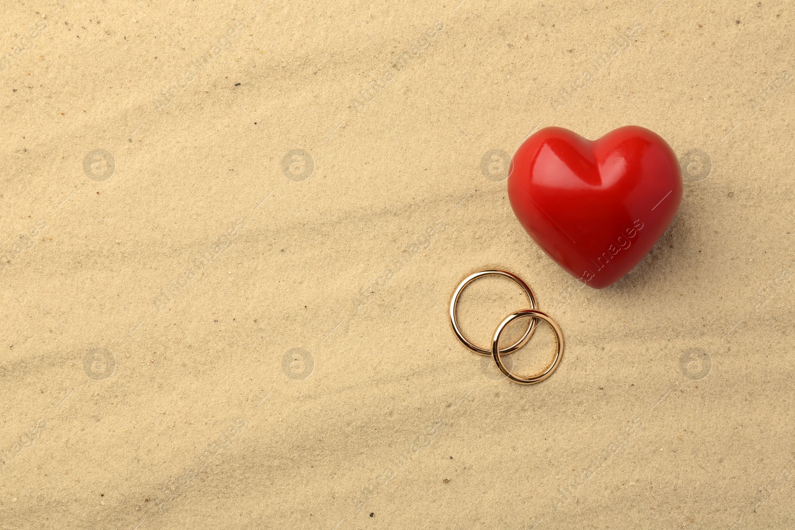 Photo of Honeymoon concept. Two golden rings and red heart on sand, top view. Space for text