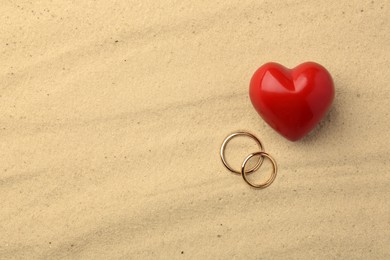 Photo of Honeymoon concept. Two golden rings and red heart on sand, top view. Space for text