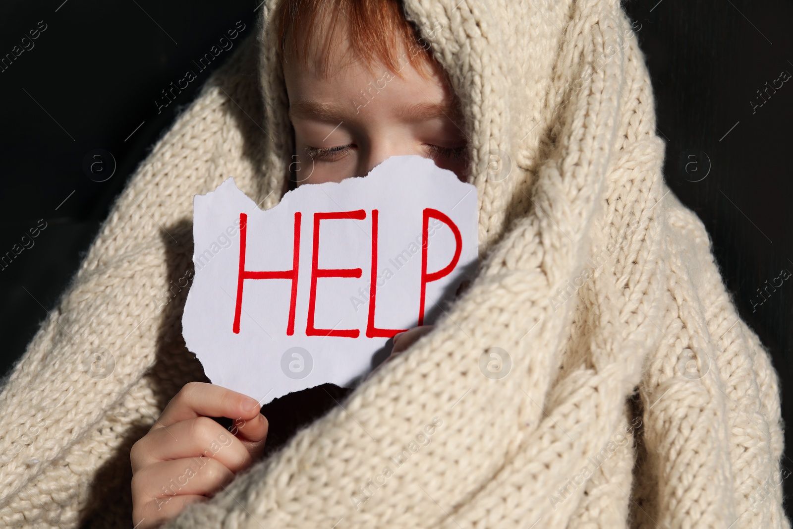 Photo of Scared little boy wrapped in blanket holding piece of paper with word Help, closeup. Domestic violence concept
