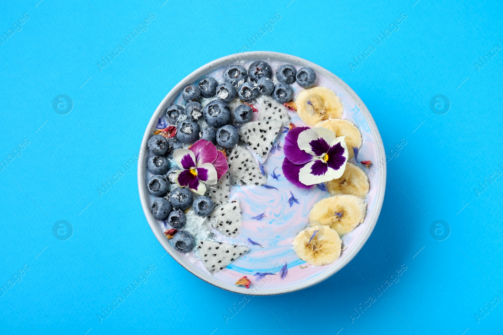 Photo of Delicious smoothie bowl with fresh fruits, blueberries and flowers on light blue background, top view