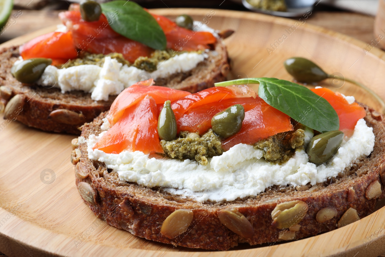 Photo of Delicious sandwiches with cream cheese, salmon, capers and pesto on wooden plate, closeup