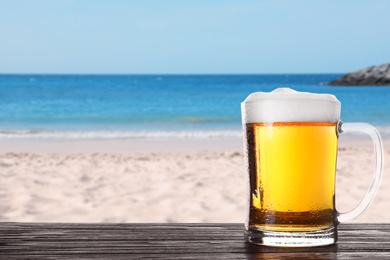 Image of Cold beer on wooden desk against blurred sea and sandy beach. Space for text