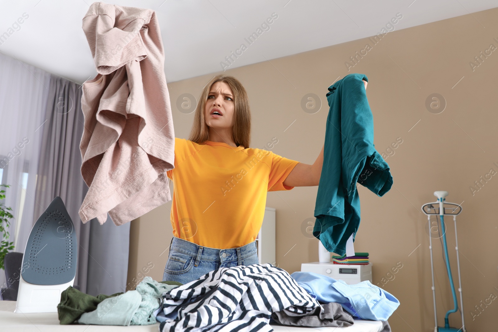 Photo of Emotional woman near board with iron and pile of clothes at home