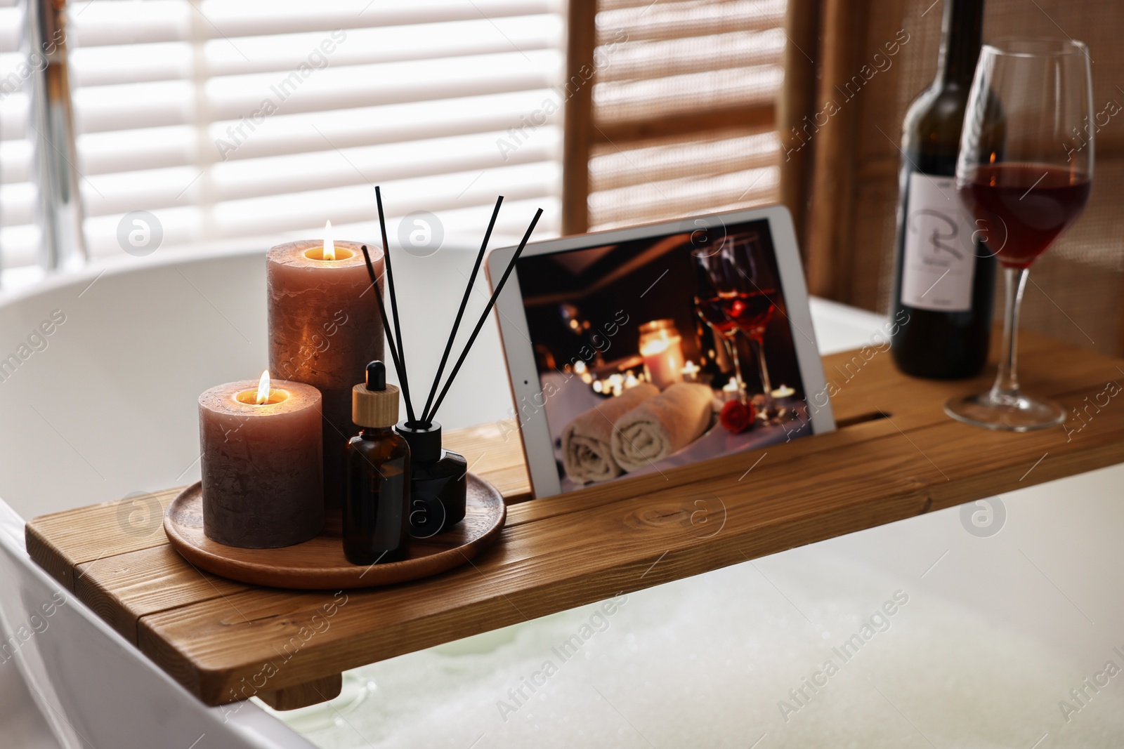 Photo of Wooden tray with tablet, burning candles and aroma products on bathtub in bathroom