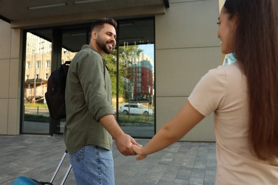 Photo of Long-distance relationship. Man with luggage holding hands with his girlfriend near building outdoors