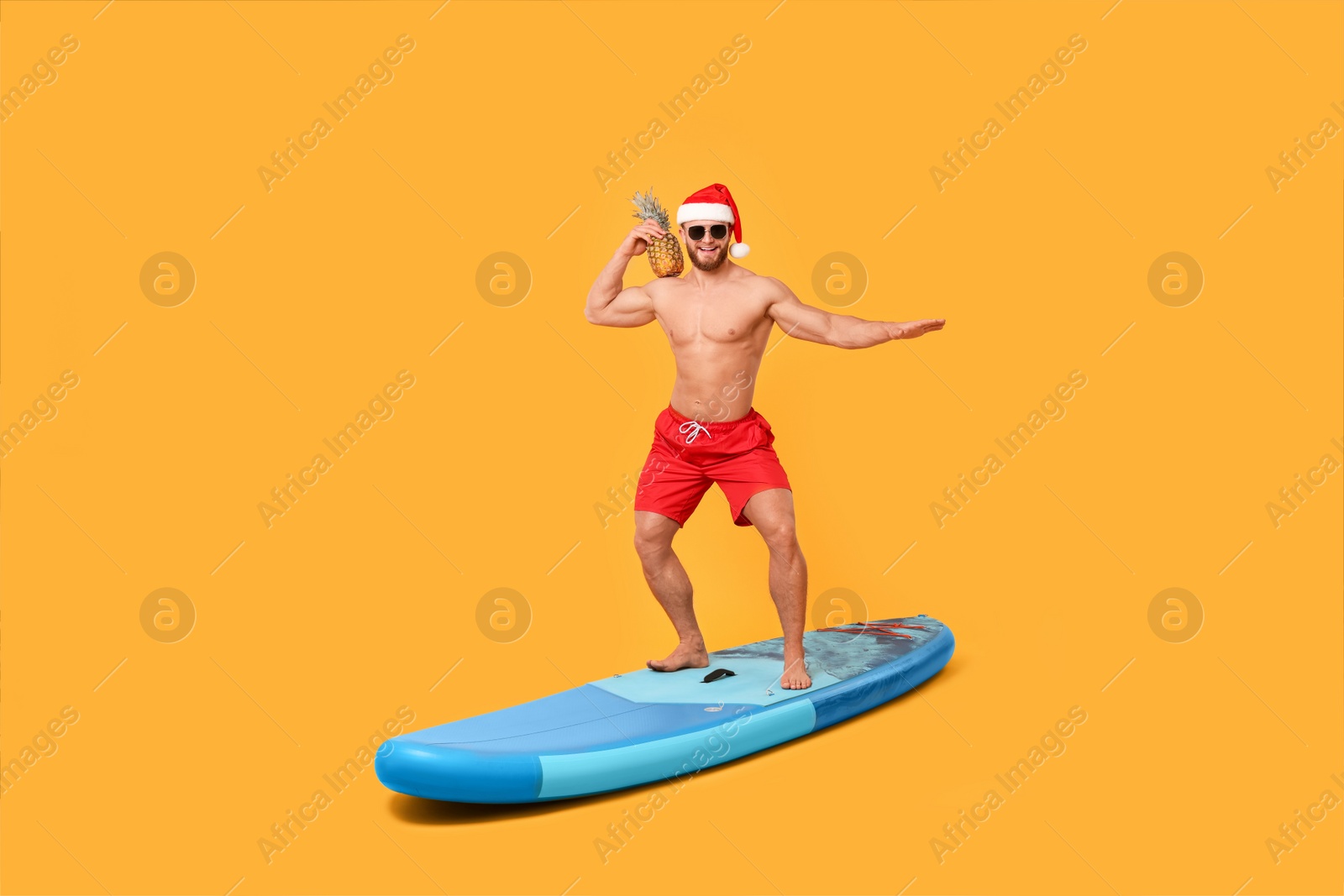 Photo of Happy man in Santa hat with pineapple posing on SUP board against orange background