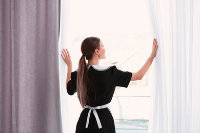 Photo of Young chambermaid opening window curtains in hotel room