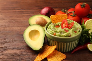 Photo of Bowl of delicious guacamole, nachos chips and ingredients on wooden table, closeup. Space for text