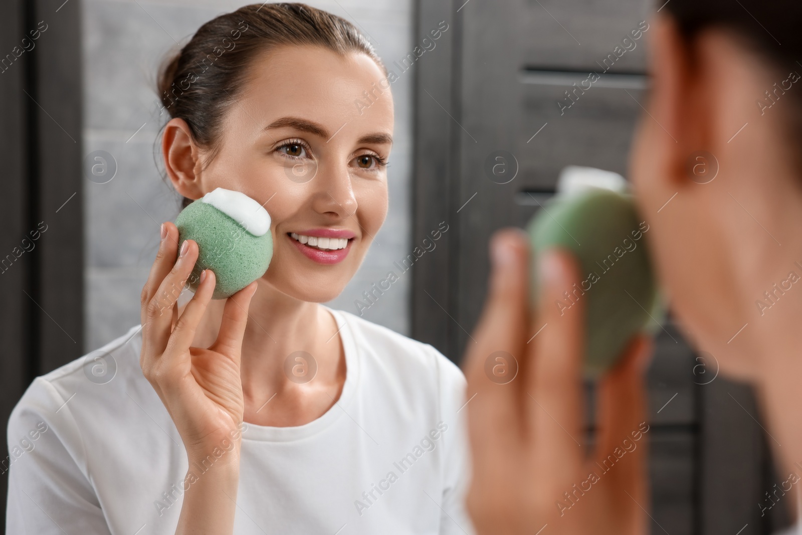 Photo of Happy young woman washing her face with sponge near mirror in bathroom