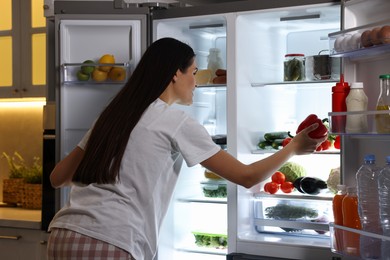 Photo of Young woman taking red bell pepper out of refrigerator in kitchen at night