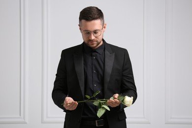 Sad man with rose flower near white wall. Funeral ceremony