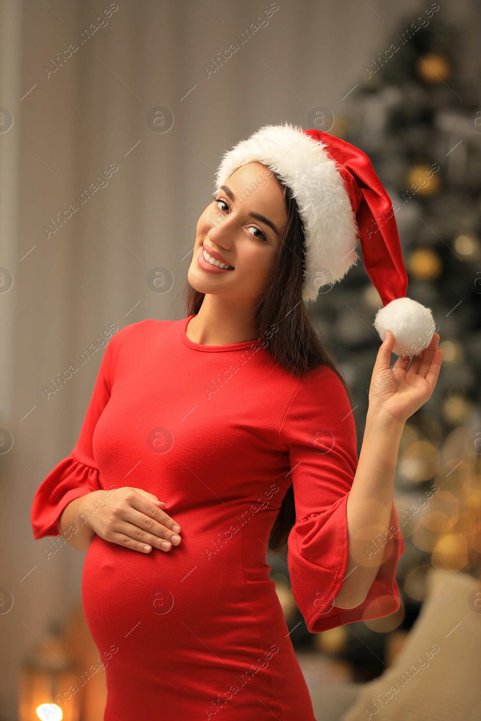 Photo of Happy pregnant woman wearing Santa hat in room decorated for Christmas. Expecting baby