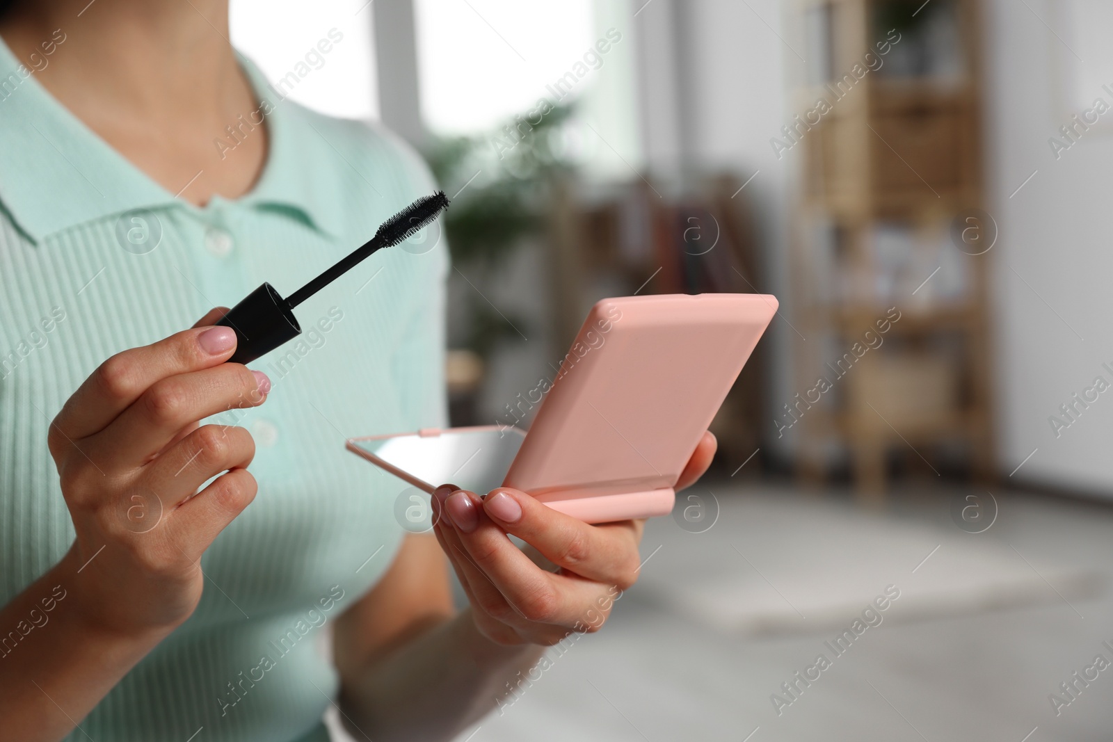 Photo of Young woman using cosmetic pocket mirror and mascara indoors, closeup