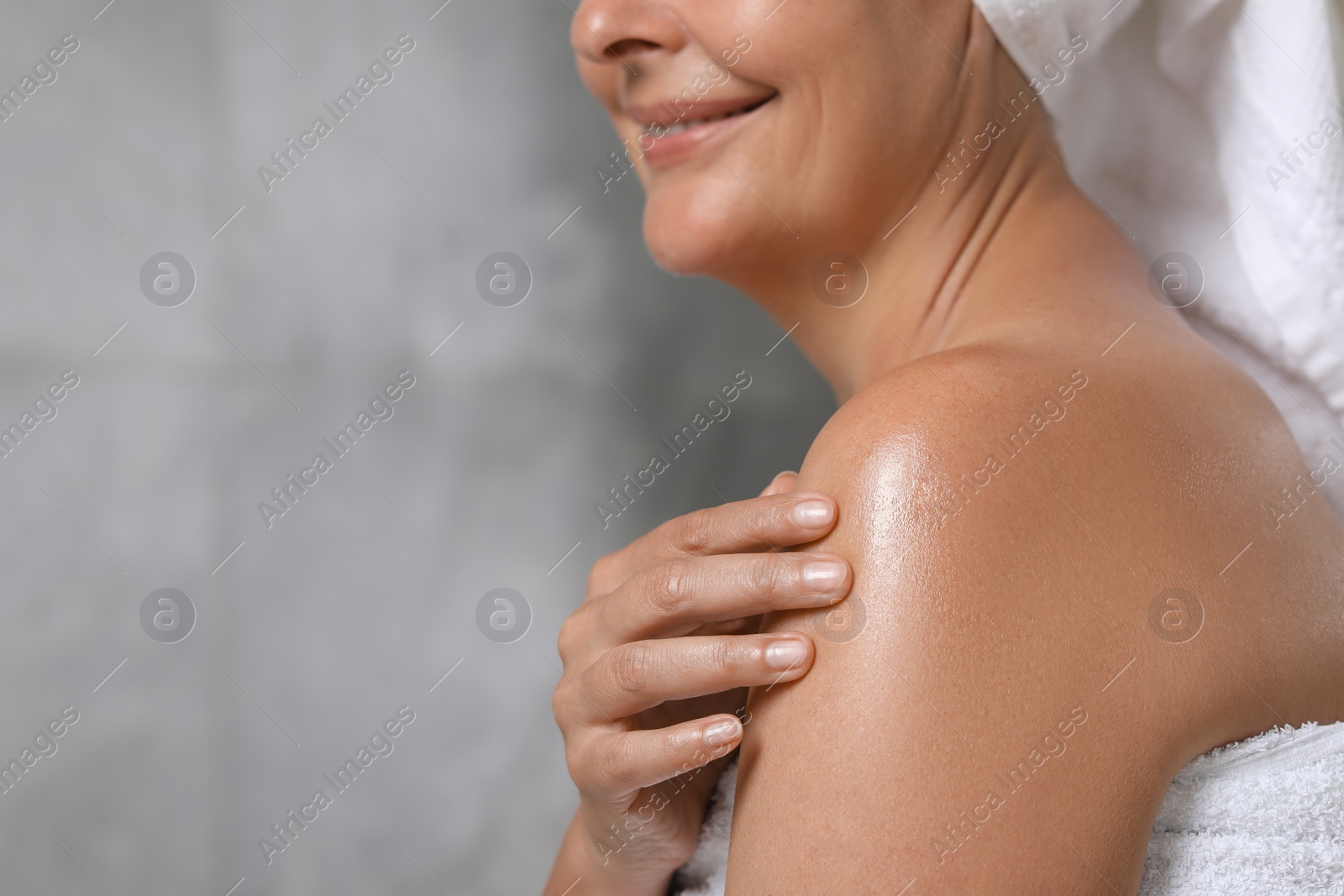 Photo of Happy woman applying body oil onto shoulder near grey wall, closeup. Space for text