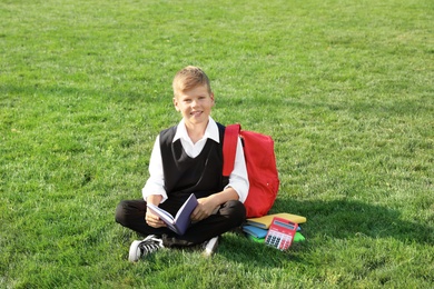 Photo of Cute boy with school stationery reading book on green lawn outdoors