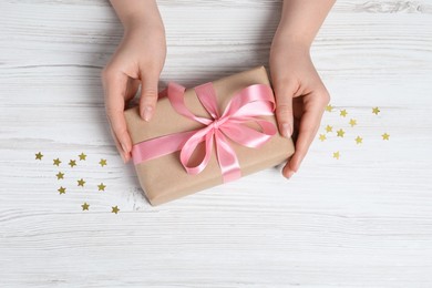 Woman holding gift box with pink bow and confetti at white wooden table, top view