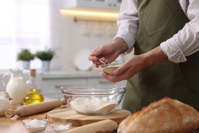 Photo of Making bread. Man putting dry yeast into bowl with flour at wooden table in kitchen, closeup