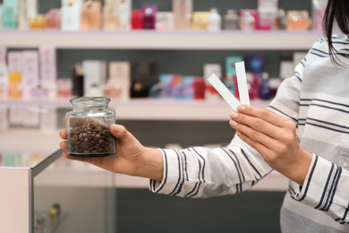 Photo of Young woman holding perfume testing stripes and jar with coffee beans in shop, closeup