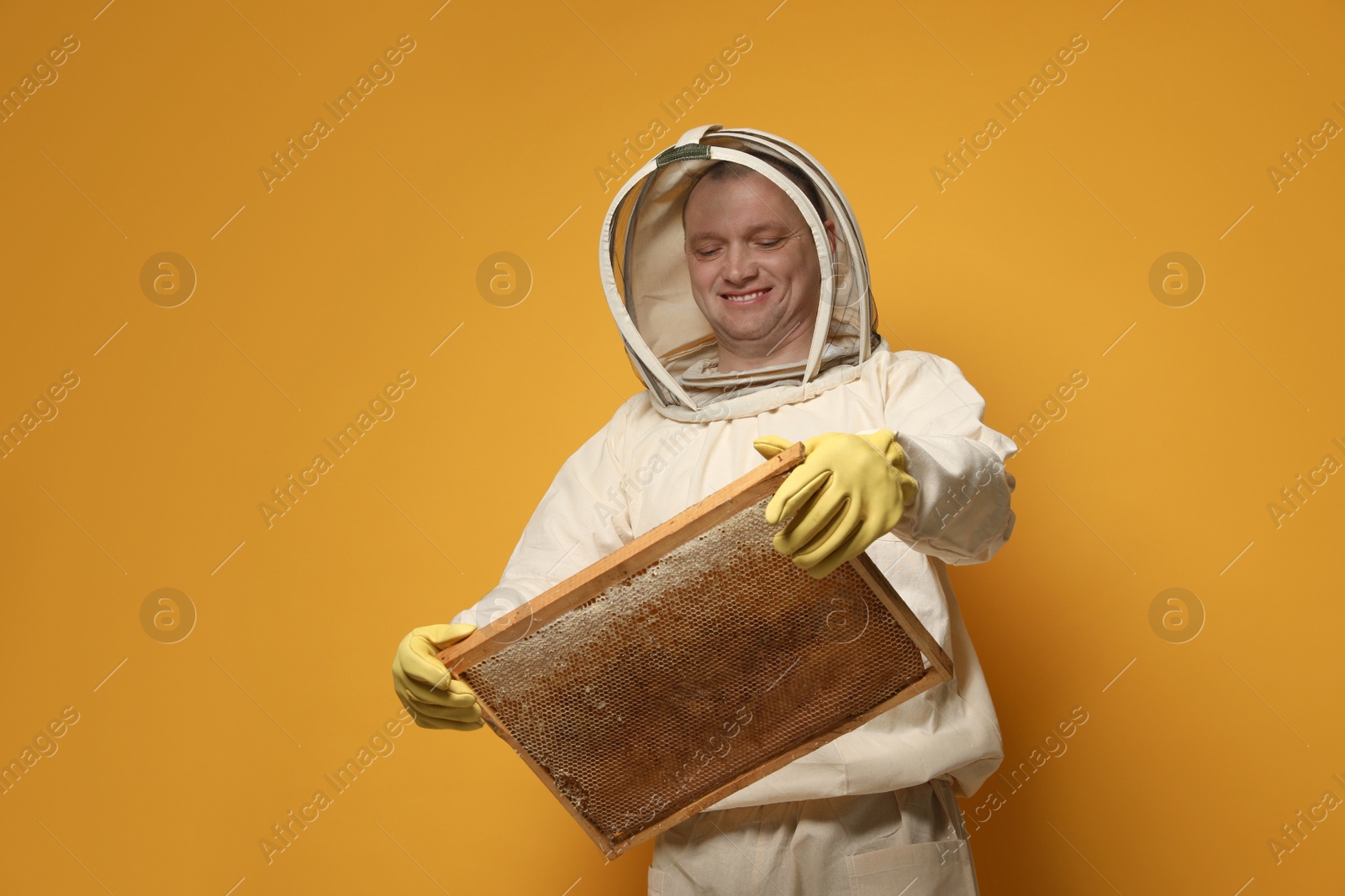 Photo of Beekeeper in uniform holding hive frame with honeycomb on yellow background