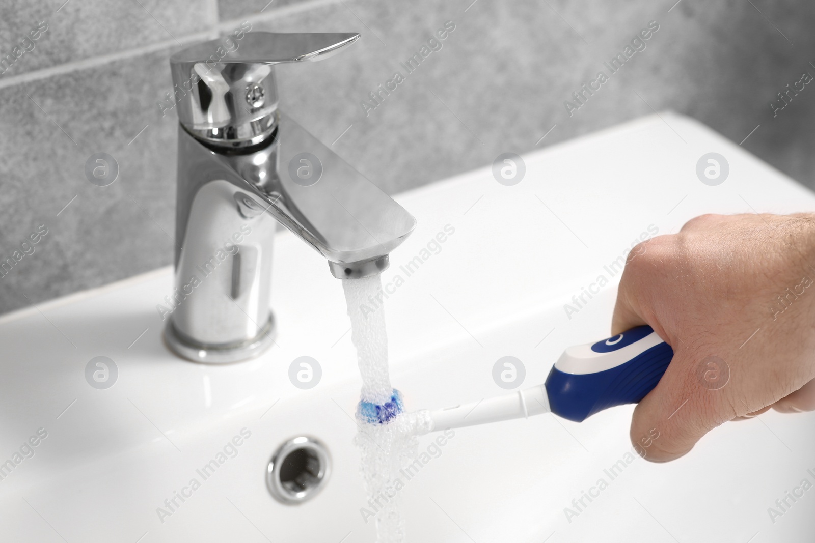 Photo of Man holding electric toothbrush under flowing water above sink in bathroom, closeup