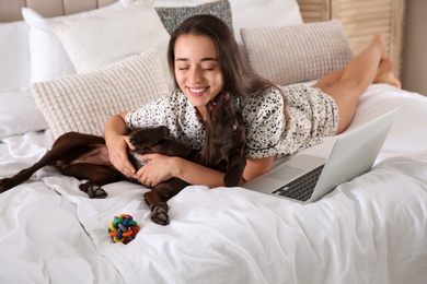 Photo of Young woman getting distracted by her dog while working with laptop in bedroom. Home office concept
