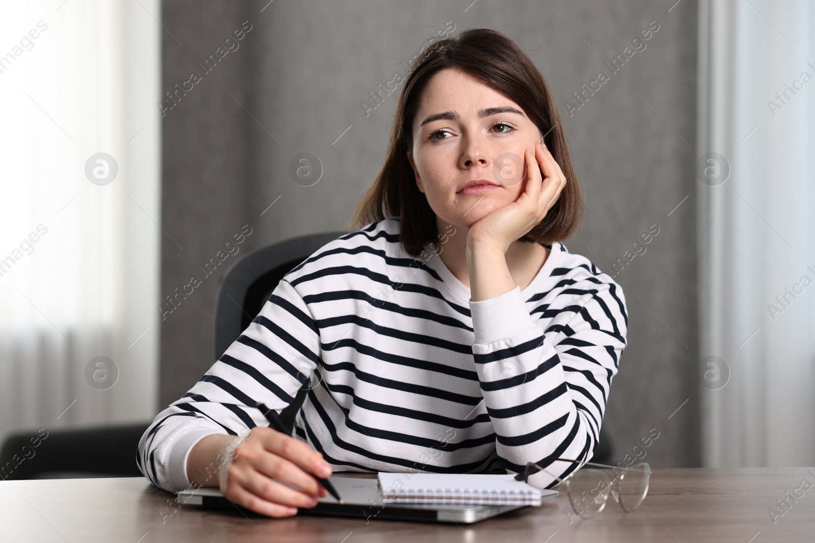 Photo of Overwhelmed woman sitting at wooden table with laptop, glasses and stationery indoors