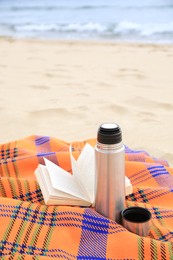 Metallic thermos with hot drink, open book and plaid on sandy beach near sea