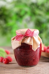 Photo of Glass jar of sweet jam with ripe raspberries on wooden table against blurred background