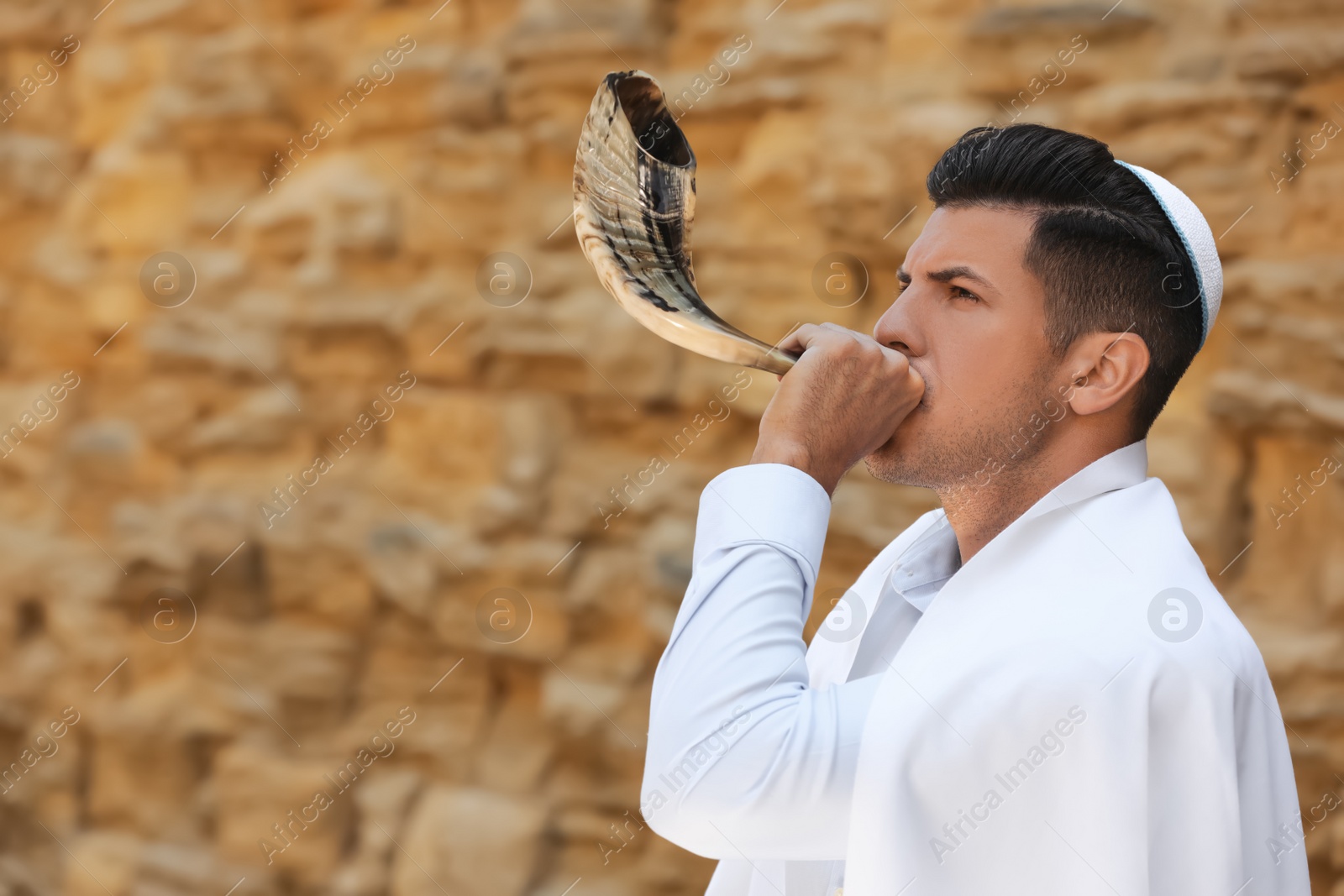 Photo of Jewish man in kippah and tallit blowing shofar outdoors. Rosh Hashanah celebration