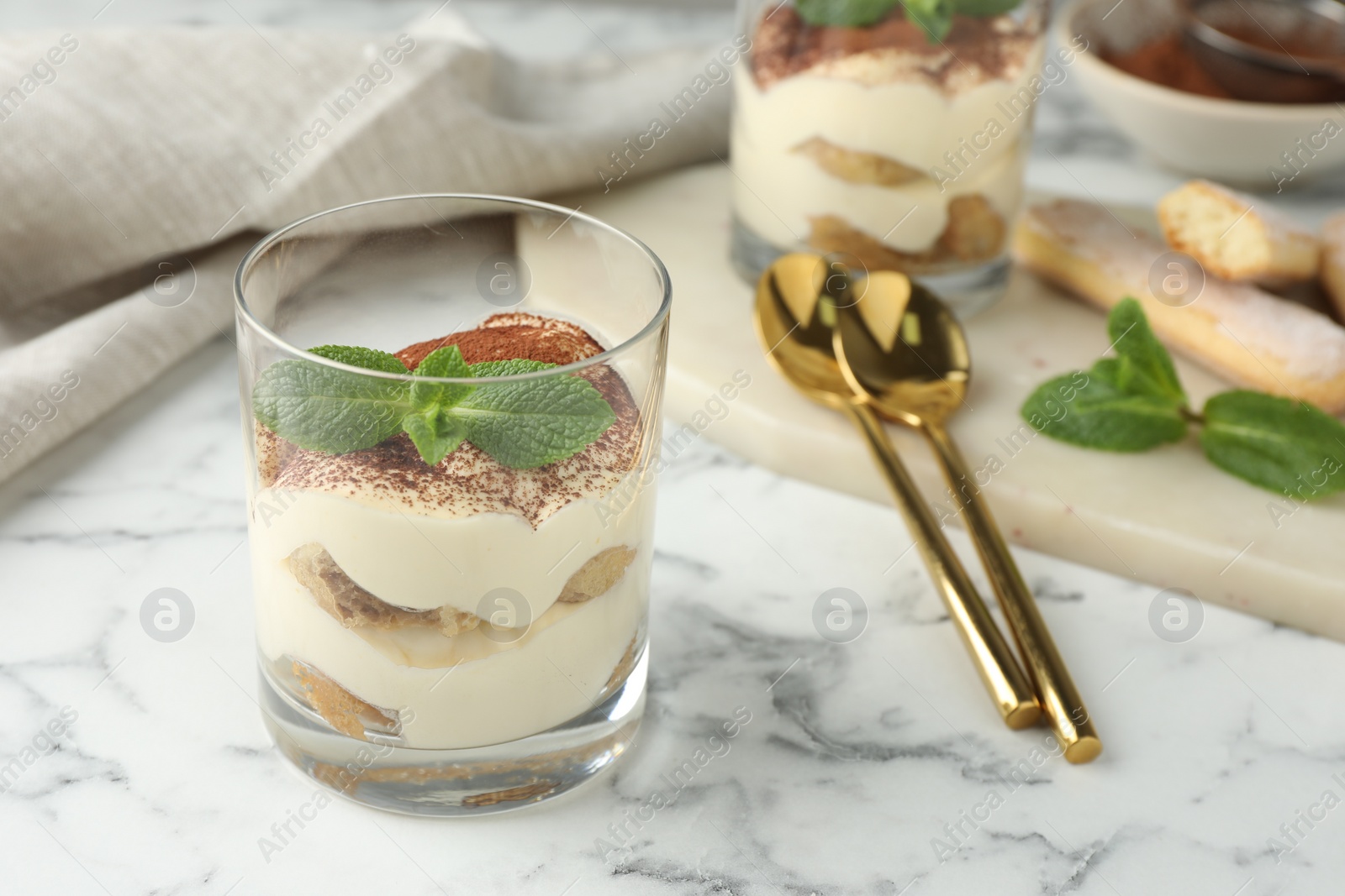 Photo of Delicious tiramisu in glass, mint leaves, spoons and cookies on white marble table