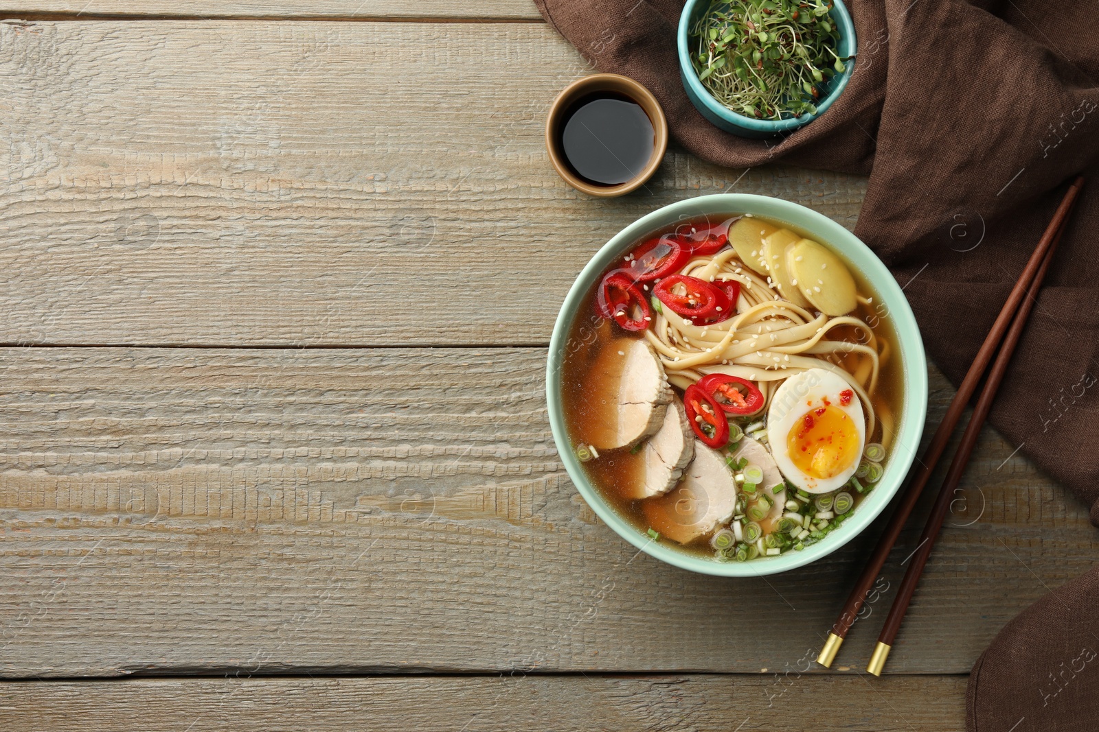 Photo of Delicious ramen in bowl, soy sauce and microgreens on wooden table, flat lay with space for text. Noodle soup