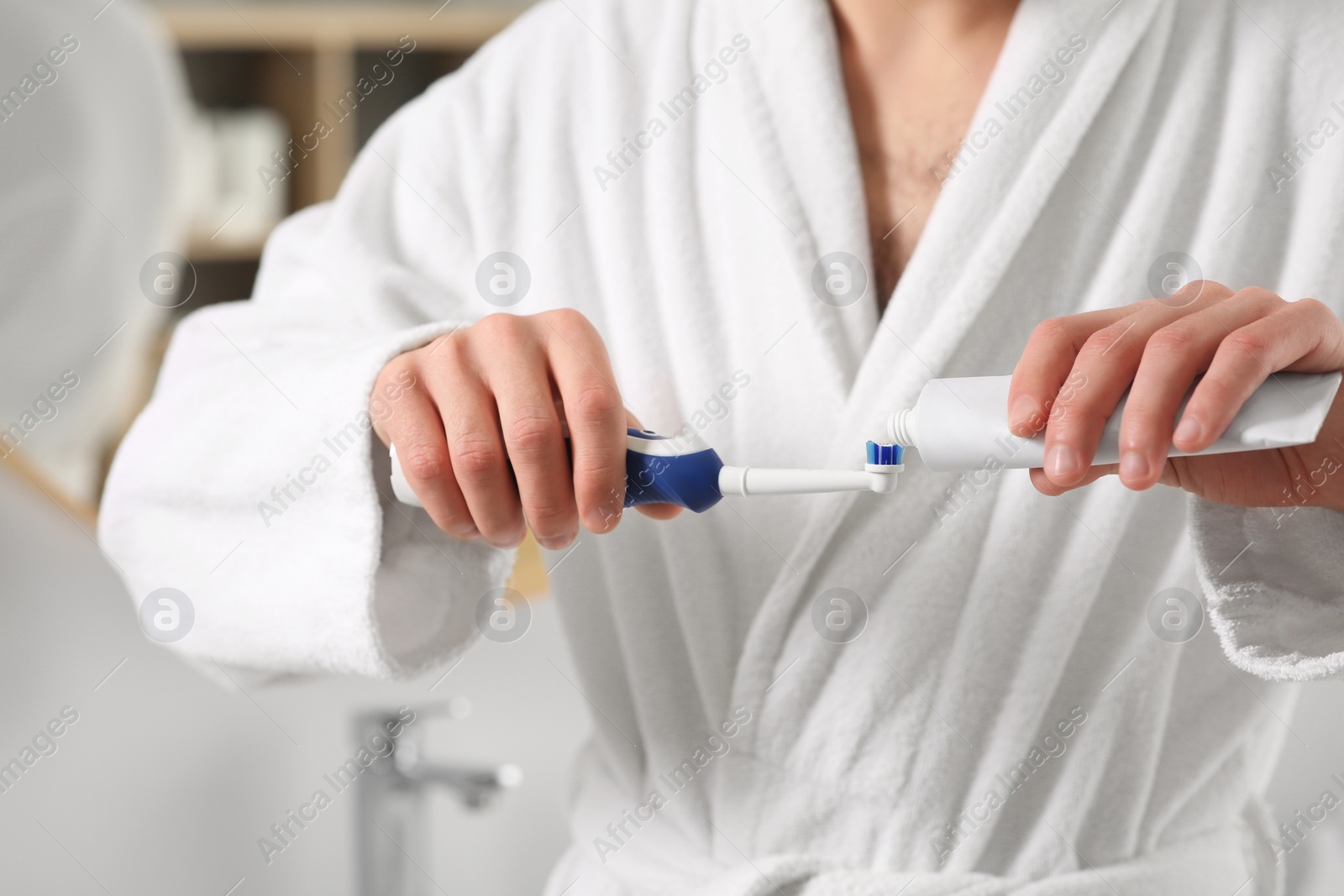 Photo of Man squeezing toothpaste from tube onto electric toothbrush indoors, closeup