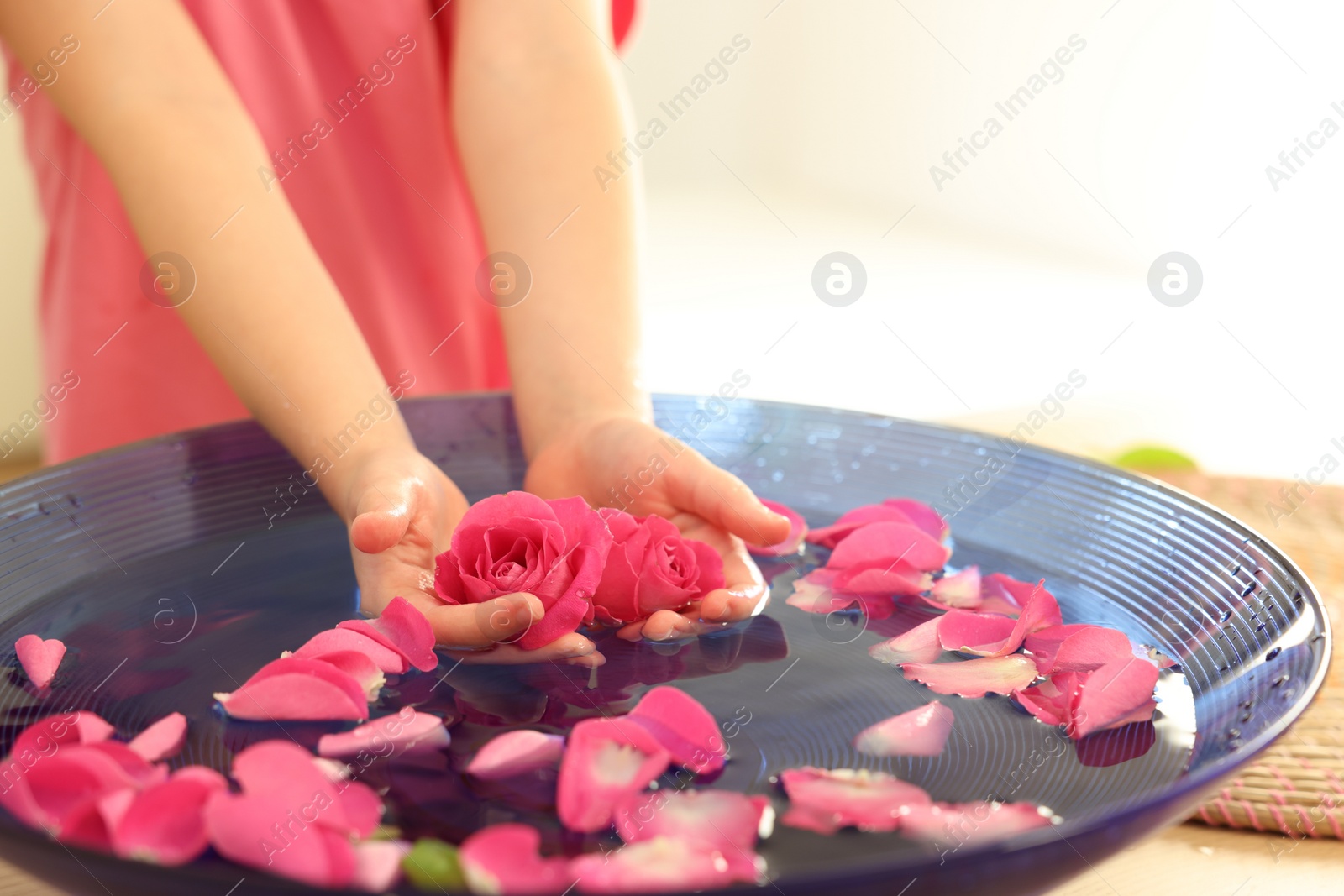 Photo of Child holding rose flowers in bowl with water and petals at table, closeup