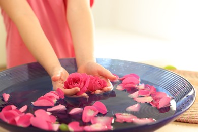 Photo of Child holding rose flowers in bowl with water and petals at table, closeup